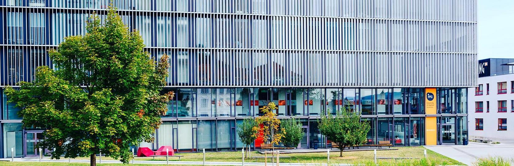 Modern office building with glass facade and horizontal slats, surrounded by trees and outdoor seating areas.