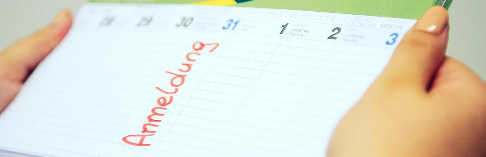 Hand hält einen offenen Kalender mit dem Wort Anmeldung in roter Schrift geschrieben.