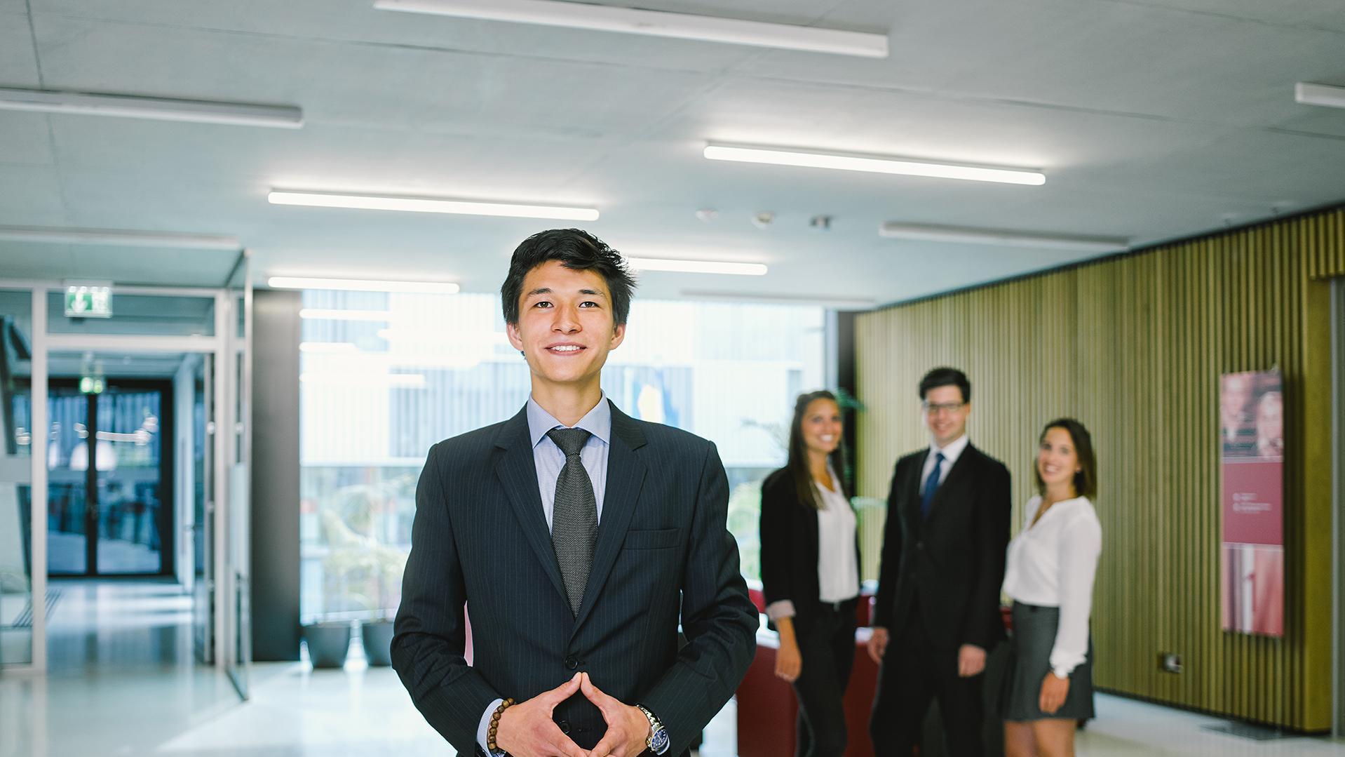Businessman with crossed hands standing in office hallway, colleagues in the background.
