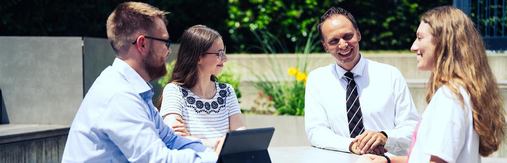 Vier Personen sitzen im Freien an einem Tisch und betrachten gemeinsam ein Laptop.