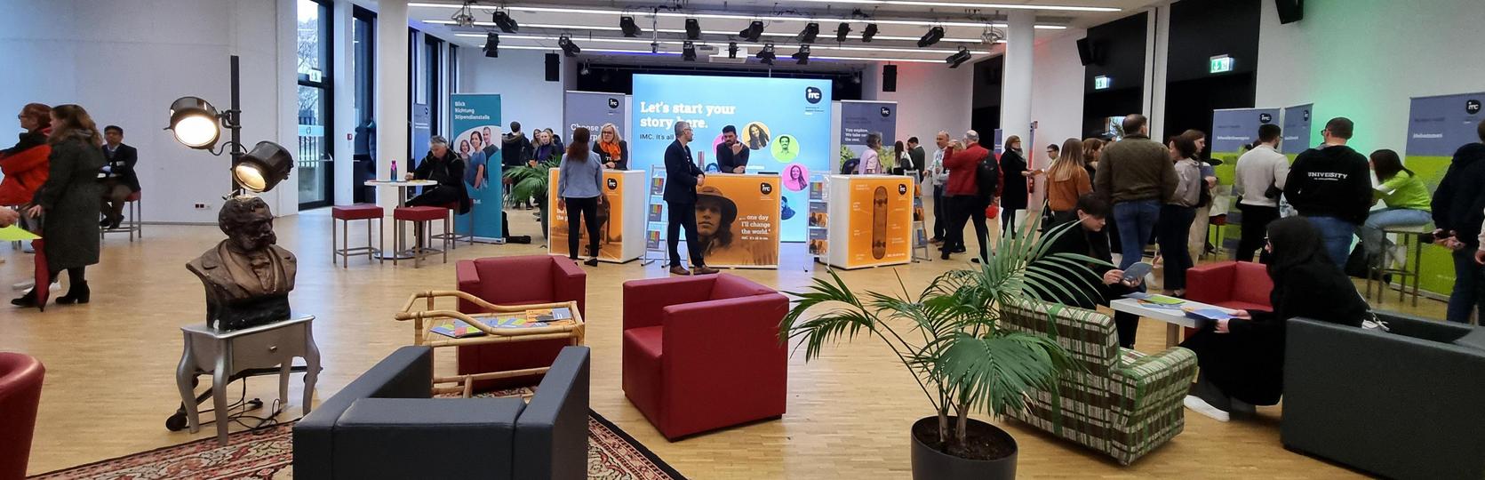 Konferenzraum mit Personen, die sich unterhalten und Informationsstände im Hintergrund.