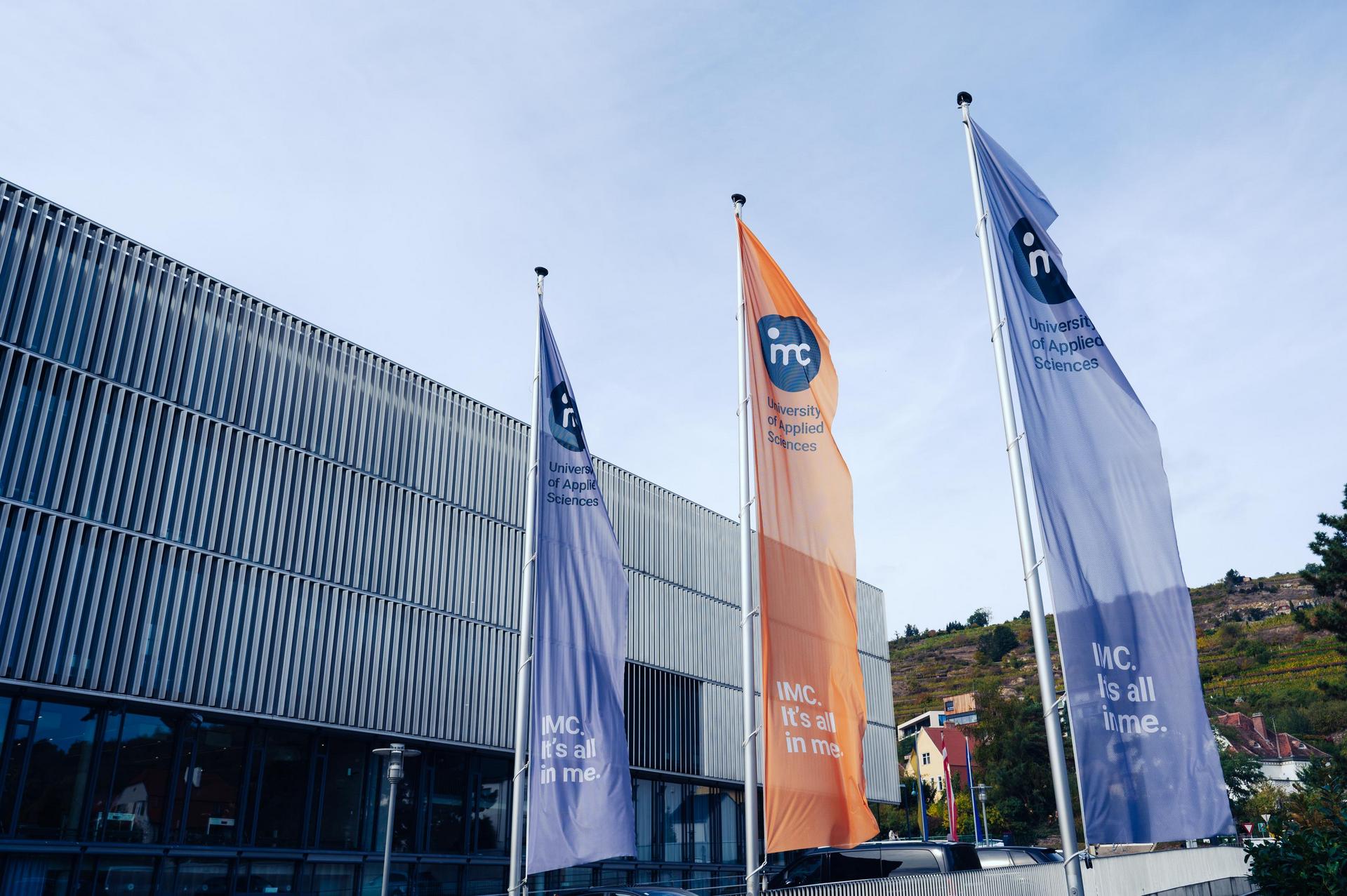 Gebäude des IMC im Hintergrund, im Vordergrund sind Flaggen mit dem Logo zu sehen