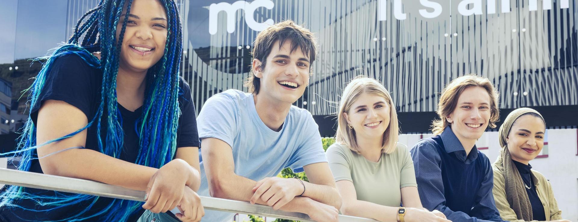Group of five young individuals leaning on a railing in front of a modern building with reflective windows and the inscription “it’s all in”.