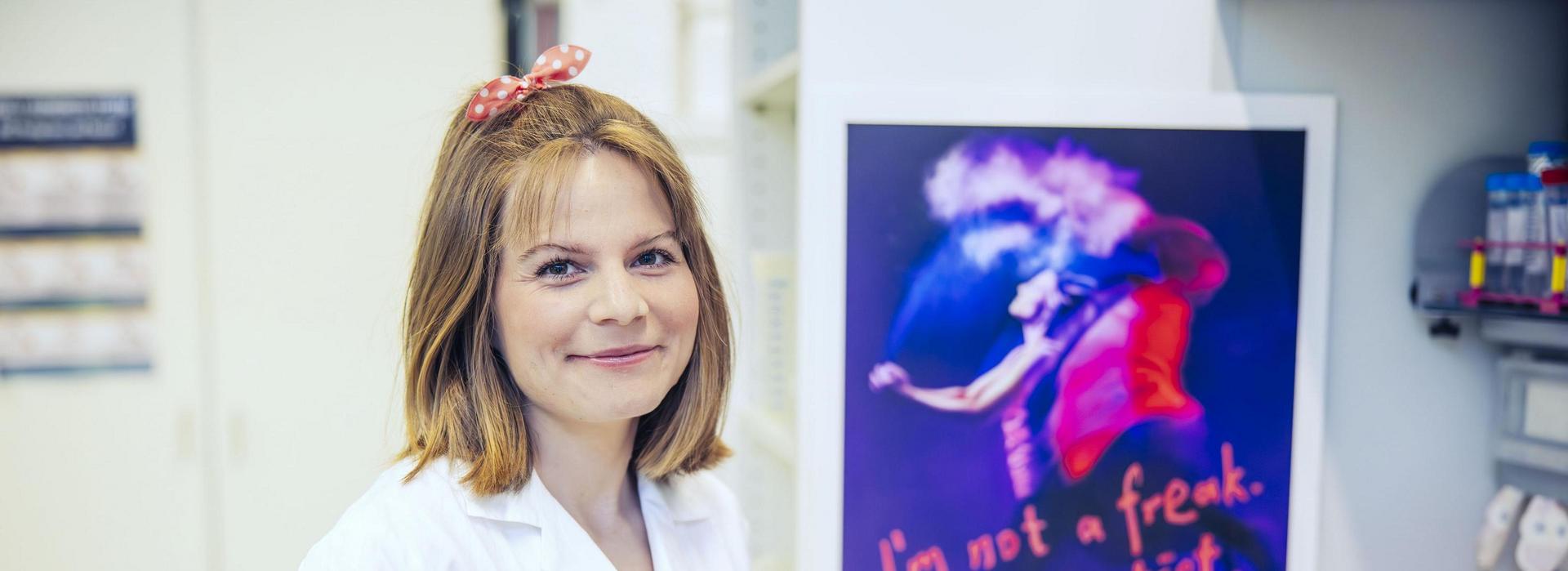 Eine Person in einem weißen Laborkittel steht vor einem Plakat mit der Aufschrift “On est fait pour s’entendre!”