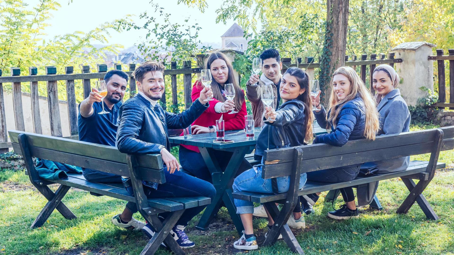 Group of people sitting outdoors at a wooden table, eating and drinking together.