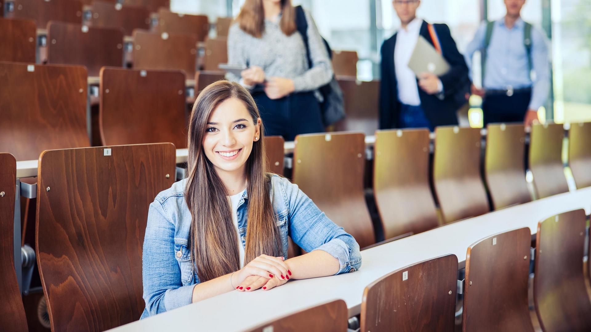 Studenten sitzen und stehen in einem Hörsaal mit Holzstühlen.
