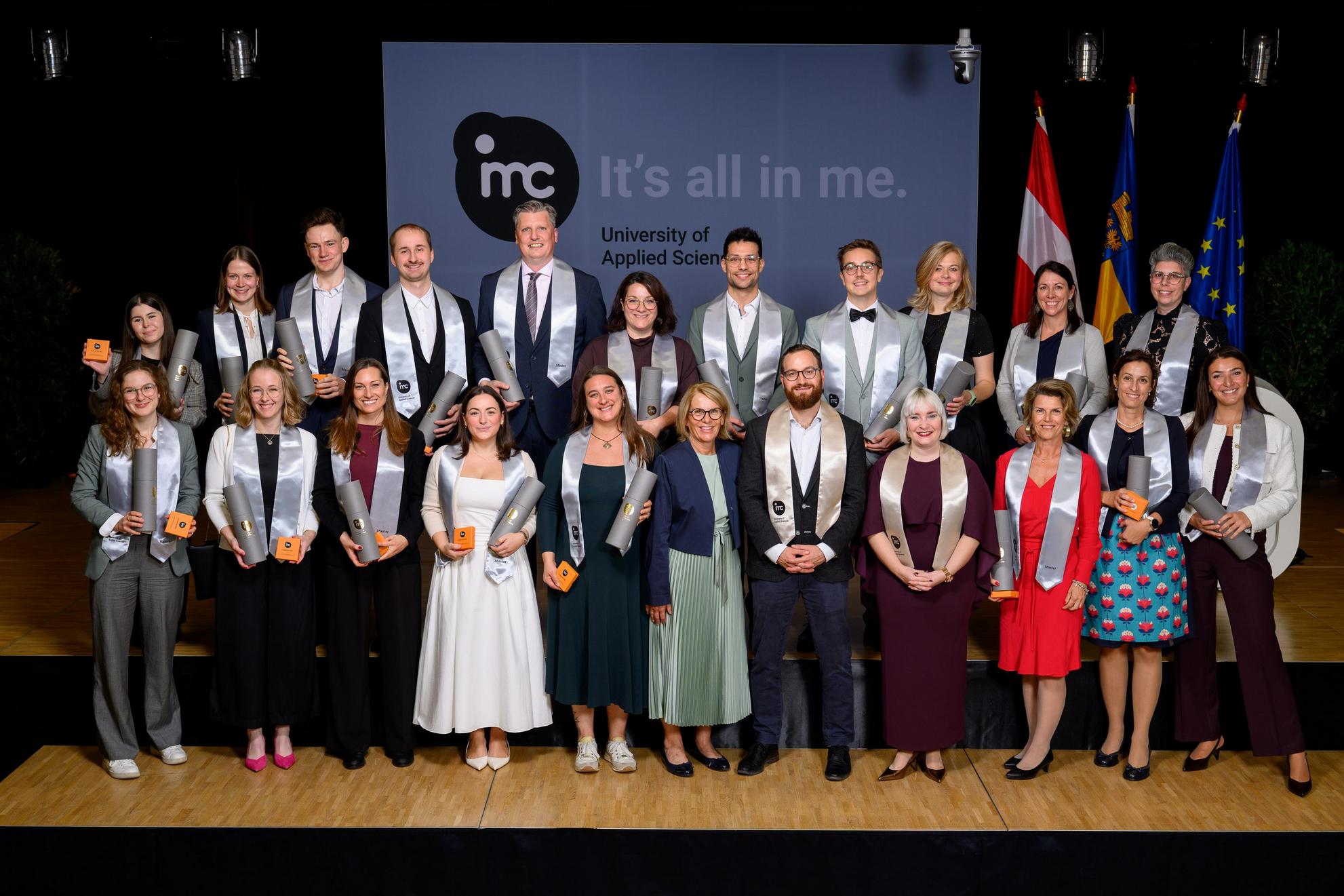 Absolventen der IMC Fachhochschule Krems mit Zeugnissen und Urkunden vor dem Banner "It's all in me."