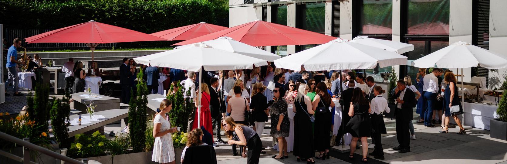 Outdoor event with people under white and red sun umbrellas in sunny weather.